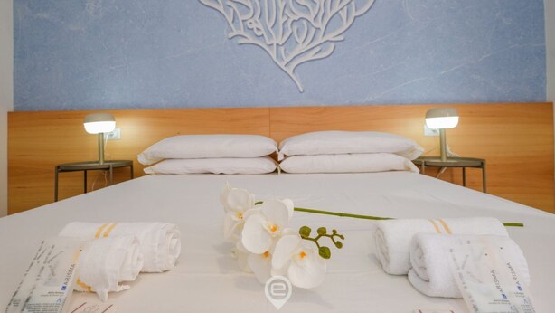 A hotel room with a neatly made bed, white towels, and a decorative wall piece. - Affittacamere Carmè - Suite Lido (Alghero)
