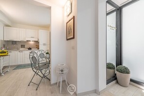 A modern kitchen with white cabinets, a dining area with a metal chair and table, and a small white stool. There are two potted plants near a glass door with a reflection.