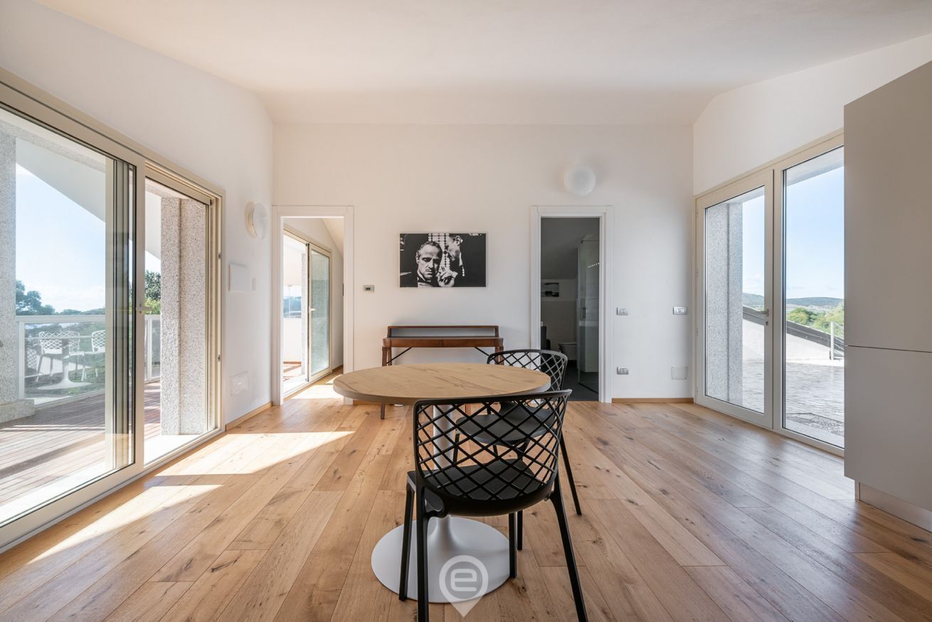 A spacious room with a wooden floor, a round table with chairs, a bench, and a large window offering a view.