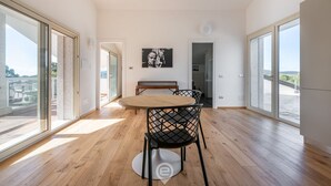 Une pièce spacieuse dotée d’un plancher en bois, d’une table ronde entourée de chaises, d’un banc et d’une grande fenêtre donnant sur un paysage agréable.