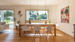 Un espace de salle à manger moderne avec une table en bois, des chaises blanches, des luminaires suspendus et une grande fenêtre donnant sur un paysage verdoyant.