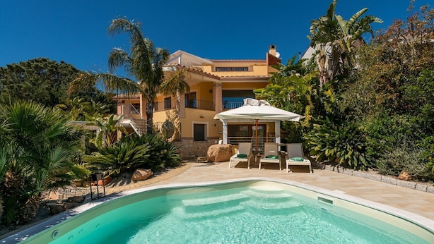 Un área de piscina con piscina azul cristalina, sillas de descanso y una zona de estar cubierta.