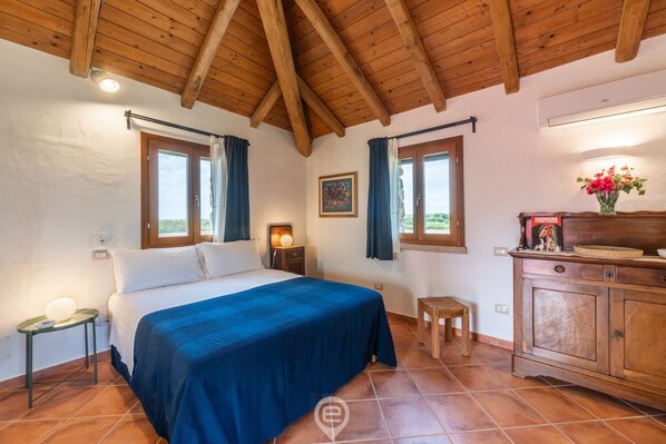 Un dormitorio con techo de madera, cama con cubrecama azul, mesita de noche, una pequeña mesa, un armario y una ventana con cortinas.