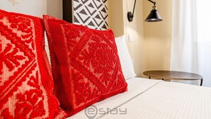 A hotel room with a bed, red patterned pillows, a geometric wall decoration, a small round table, and a wall-mounted lamp.