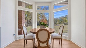 Un coin repas avec une table ronde et quatre chaises, de grandes fenêtres donnant sur des arbres et un mur en briques, et une plante en pot posée sur la table.