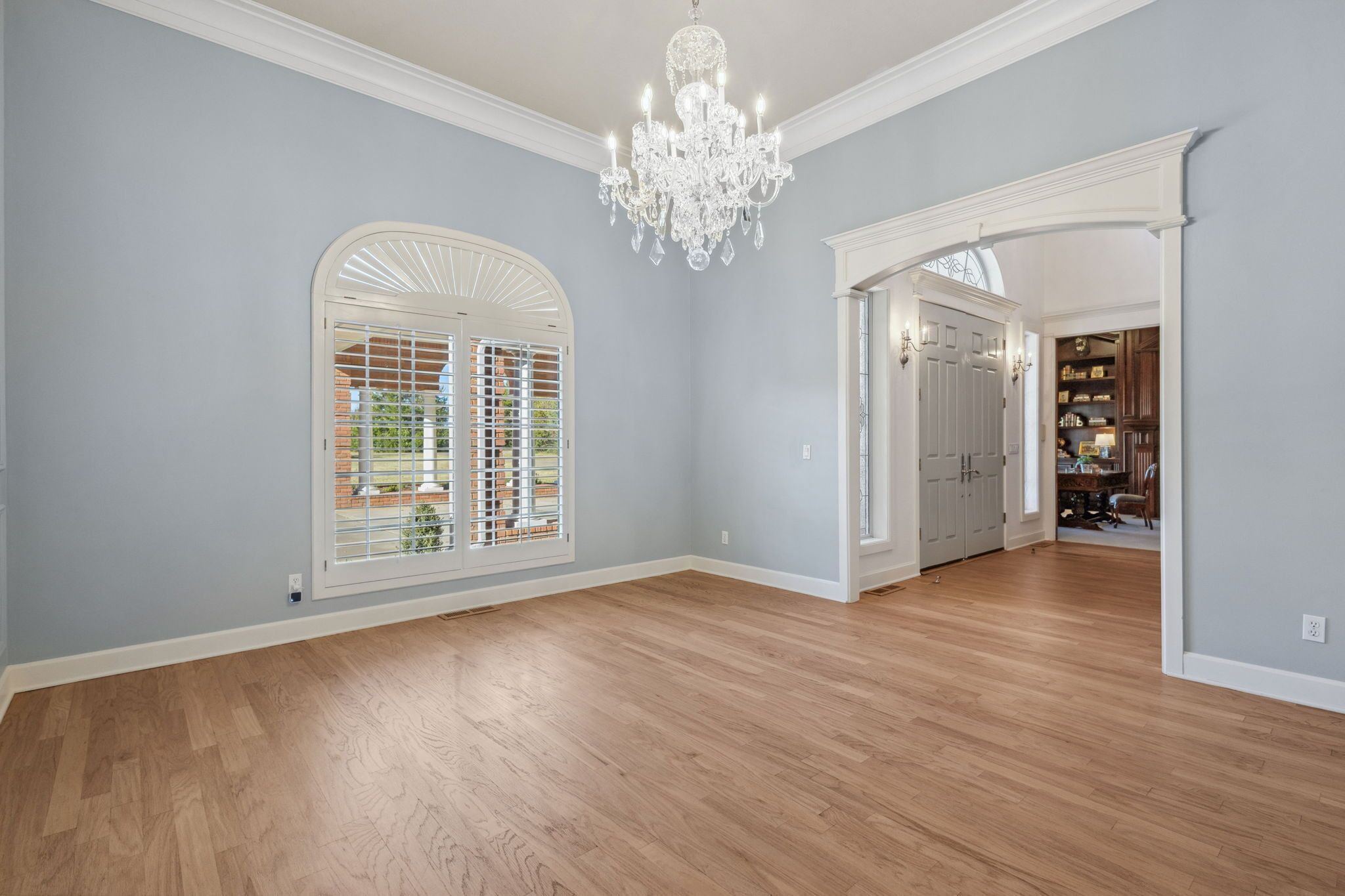 Una habitación amplia con piso de madera, un gran ventanal arqueado con persianas y un candelabro.