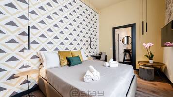 A modern hotel room with a geometric-patterned wall, a neatly made bed with towels, a small round table, and a potted plant.