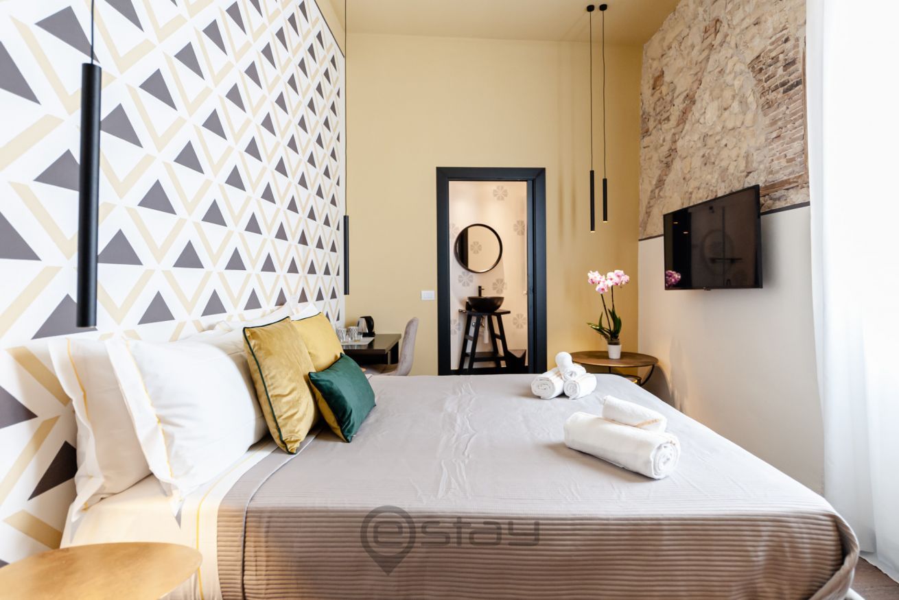 Een moderne hotelkamer met een wand met een geometrisch patroon, een bed met opgerolde handdoeken, een bijzettafeltje met een plant en een aan de wand bevestigde televisie.
