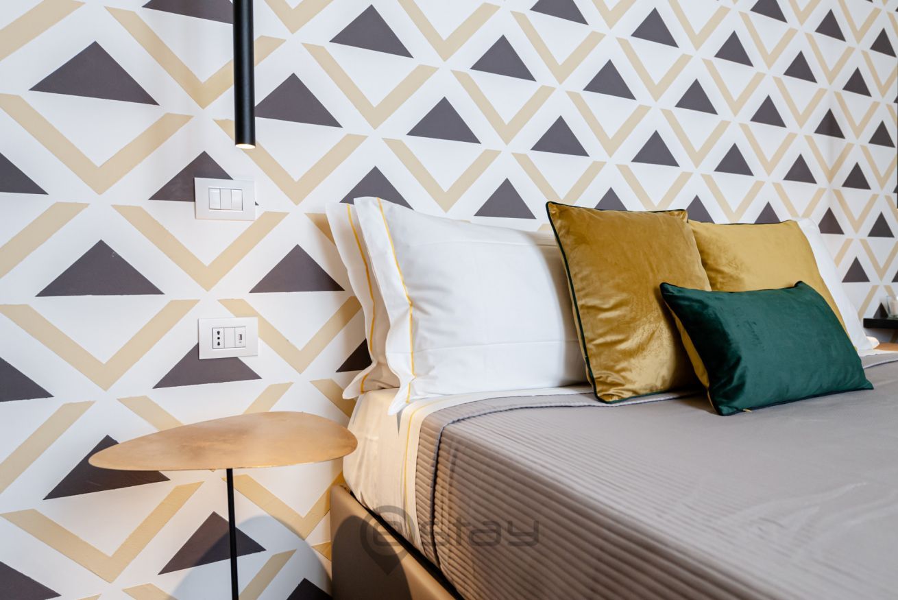 Una camera d'albergo ordinata con carta da parati decorata, un letto con cuscini, un comodino e una lampada.