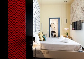 A modern hotel room with a bed, a TV, a small table with a plant, and a decorative wall. - KyS Castello - Suite Villanova (Cagliari)