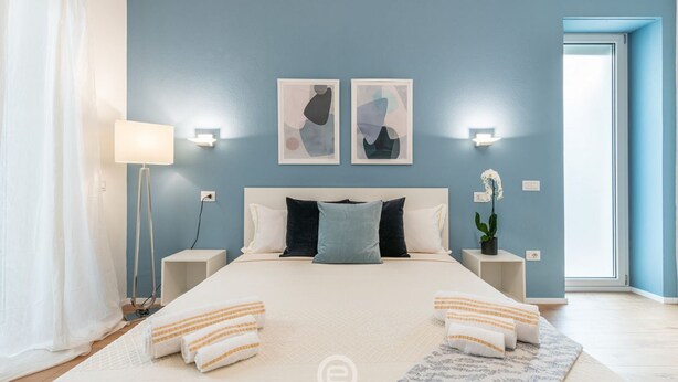 Una camera da letto con un letto grande, due lampade a parete, un comodino con lampada, una pianta in vaso e una finestra con tende.