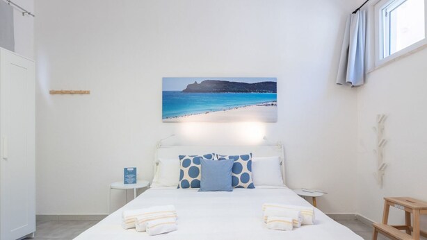 A hotel room with a bed, a bedside table, a nightstand, a window with curtains, and a wall-mounted picture of a beach. - Saturnino - Apartment (Cagliari)