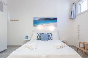 A hotel room with a bed, a bedside table, a nightstand, a window with curtains, and a wall-mounted picture of a beach.