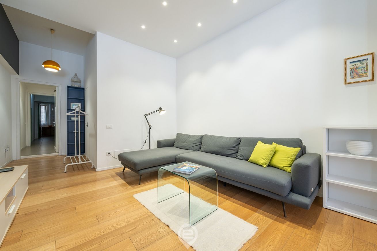 A modern living room with a grey sectional sofa, a glass coffee table, and a white rug. There is a white shelving unit to the right and a doorway leading to another room.