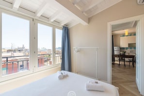 Une chambre d’hôtel avec un lit, une fenêtre avec des rideaux, une vue sur la ville et une kitchenette dans la pièce adjacente.