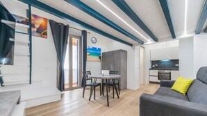 Un salon moderne avec un canapé gris, une table à manger avec des chaises, un coin cuisine et un escalier offrant une vue sur l’extérieur.