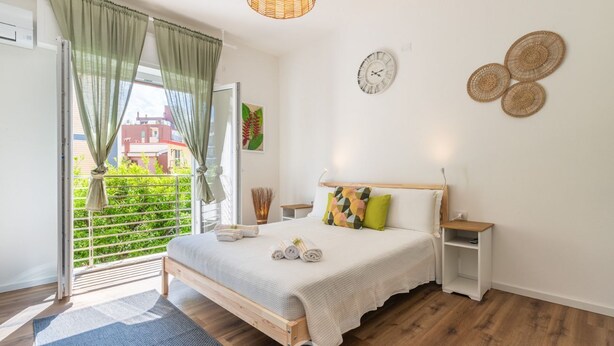 A bedroom with a wooden bed, a bedside table, a nightstand, a window with curtains, a wall clock, and a view of greenery outside.