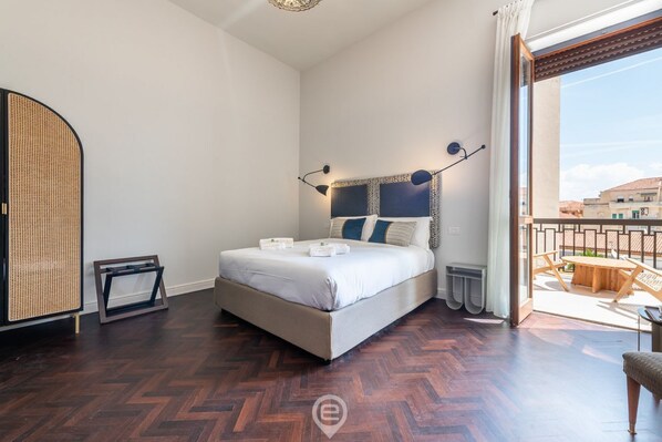 Une chambre avec un grand lit, une chaise en rotin, une table en bois et un balcon donnant sur des immeubles.