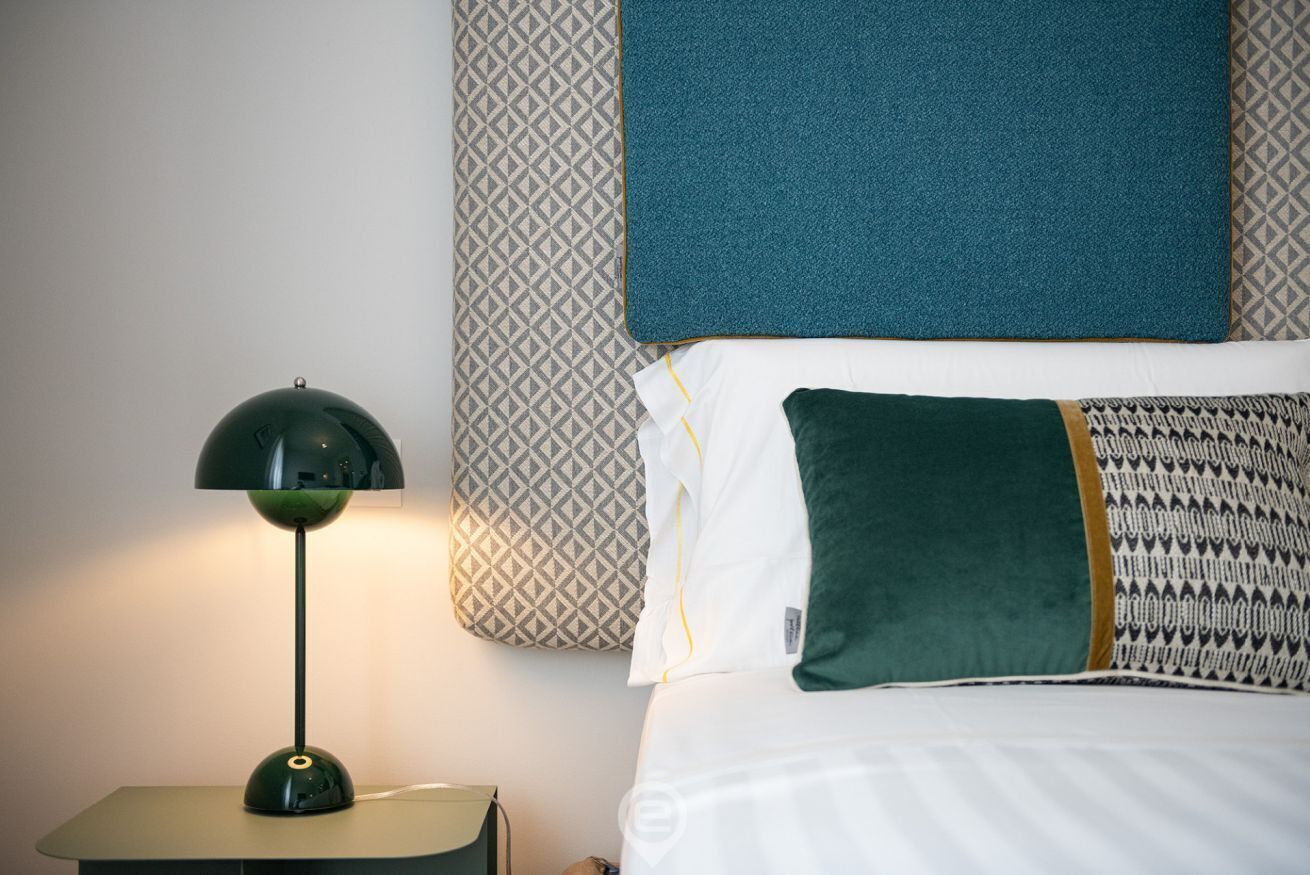 A neatly made bed with patterned pillows, a green lamp on a bedside table, and a patterned wall behind the bed.