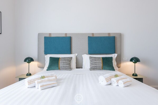 A neatly made bed with white linens and striped towels, two bedside tables with lamps, and patterned pillows. - Verso Suites - Suite Turchese (Cagliari)