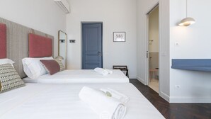 A hotel room with two beds, a blue door, a mirror, a framed picture, and a bedside table with a lamp. - Verso Suites - Suite Quarzo (Cagliari)