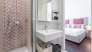 A modern bathroom with a shower, a sink, and a mirror. There is a bed with a headboard and pillows in the adjacent room. - Verso Suites - Suite Quarzo (Cagliari)