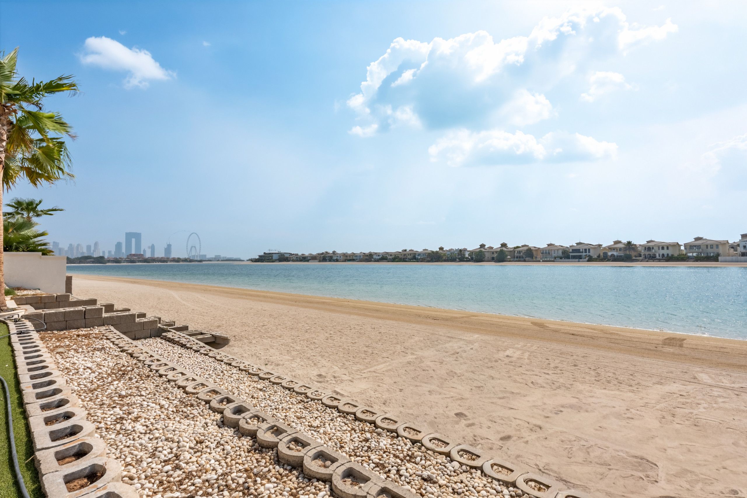Een strand met een heldere lucht, een rustige zee en een rij huizen in de verte.