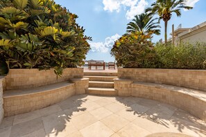 Un jardin en contrebas, offrant une vue sur la mer, entouré d’une végétation tropicale luxuriante et agrémenté d’un banc en pierre.