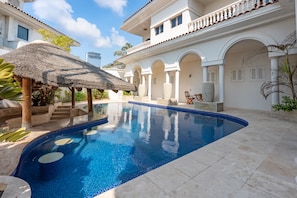 A swimming pool with a thatched roof structure, surrounded by a white building with arched walkways and a tiled deck.