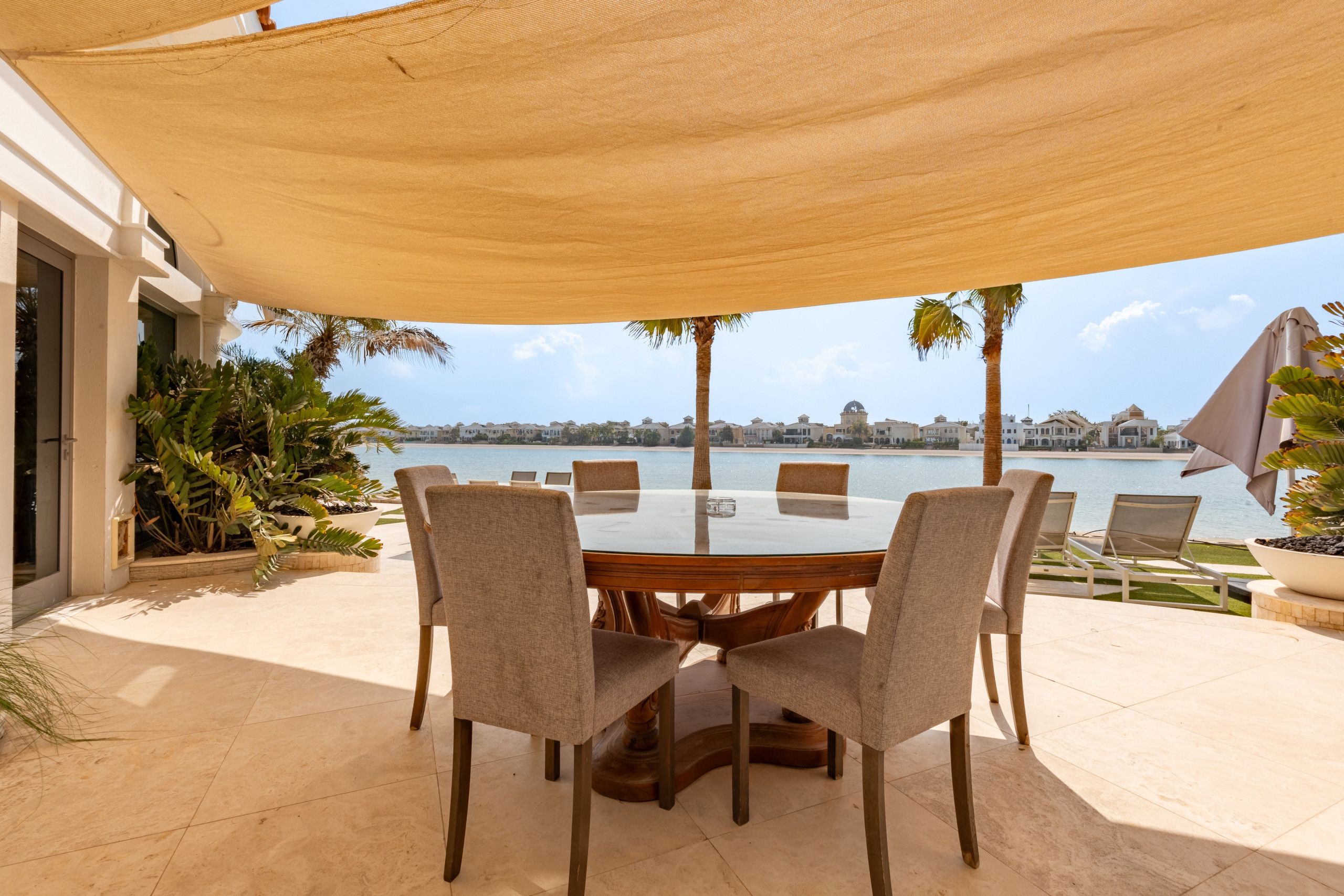 Een buiten eethoek met een ronde tafel en stoelen, een grote parasol, en uitzicht op een waterpartij en gebouwen op de achtergrond.