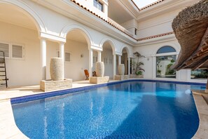 Una piscina con borde de azulejos, rodeada de columnas y arcos blancos, y vista a un edificio con techo de tejas.