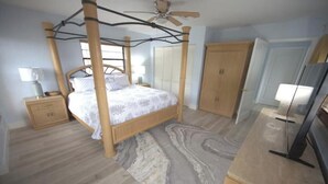 A four-poster bed with a wooden frame and white bedding, a wooden nightstand with a lamp, a wardrobe, and a television on a wooden cabinet.