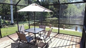A covered outdoor patio with a table and chairs, overlooking a pond and greenery.