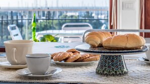 Une table à manger recouverte d’une nappe blanche, une assiette de biscuits, une tasse blanche et un présentoir à gâteaux bleu et blanc, sur lequel reposent deux croissants.