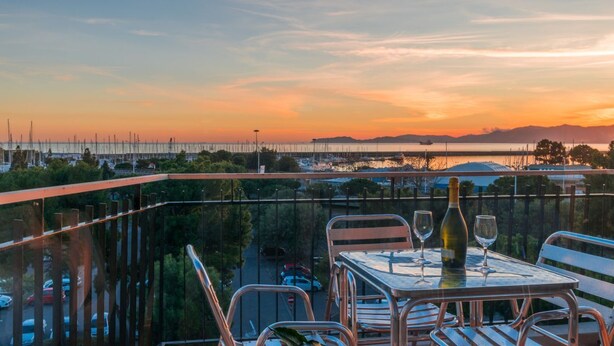 Un balcon avec une table dressée pour deux personnes, donnant sur une marina avec des bateaux et offrant une vue sur le coucher du soleil.