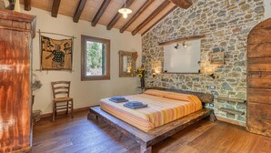 A rustic bedroom with a stone wall, wooden beams, a bed with striped bedding, a wooden bench, a chair, and a wooden wardrobe. - 3 bedroom pet friendly home in Imperia (Imperia)