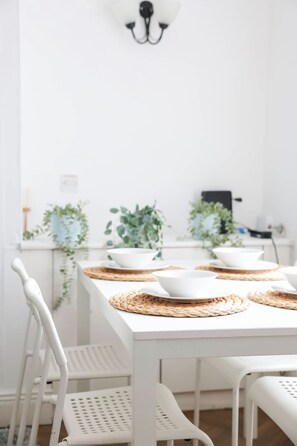 Taula de dinar blanca amb sotsplat, cadires blanques i plantes verdes al fons.
