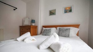 Un lit bien fait, avec du linge de lit blanc et des serviettes pliées, une table de chevet en bois avec une lampe, et deux tableaux encadrés accrochés au mur.