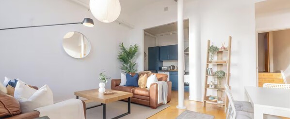 Un salon moderne avec un canapé, une table basse et un coin repas. On aperçoit une cuisine à l’arrière-plan, avec des meubles bleus.