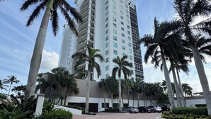 Um hotel alto com vários andares, cercado por palmeiras e um caminho de entrada pavimentado.