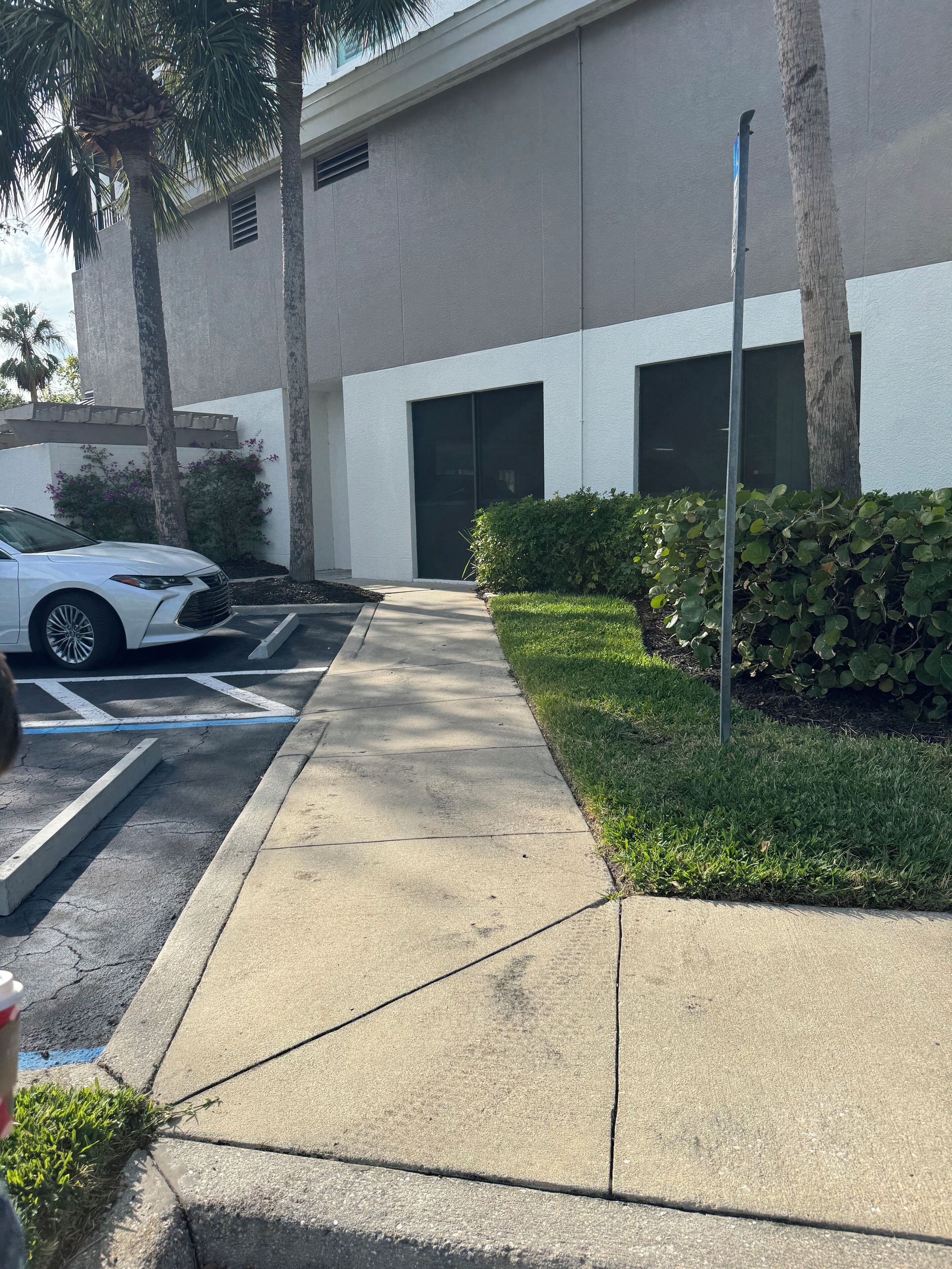 A parking area with a white car, a sidewalk, and a building with a large window and a door.