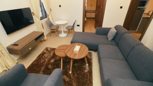 A modern living room with a grey sofa, a flat-screen TV, a small round table, and a brown furry rug. - AJs Serenity Suites (Abuja)