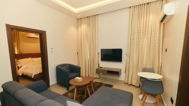 A modern hotel room with a large grey sofa, a smaller grey armchair, a round coffee table, a flat-screen TV mounted on the wall, and a small round table with two chairs. - AJs Serenity Suites (Abuja)