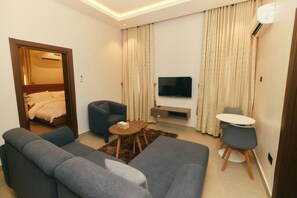 A modern hotel room with a large grey sofa, a smaller grey armchair, a round coffee table, a flat-screen TV mounted on the wall, and a small round table with two chairs. - AJs Serenity Suites (Abuja)