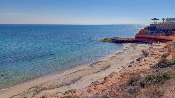 Une plage côtière aux eaux bleues et limpides, avec un rivage sablonneux et une construction dotée d’un toit, située sur la droite.