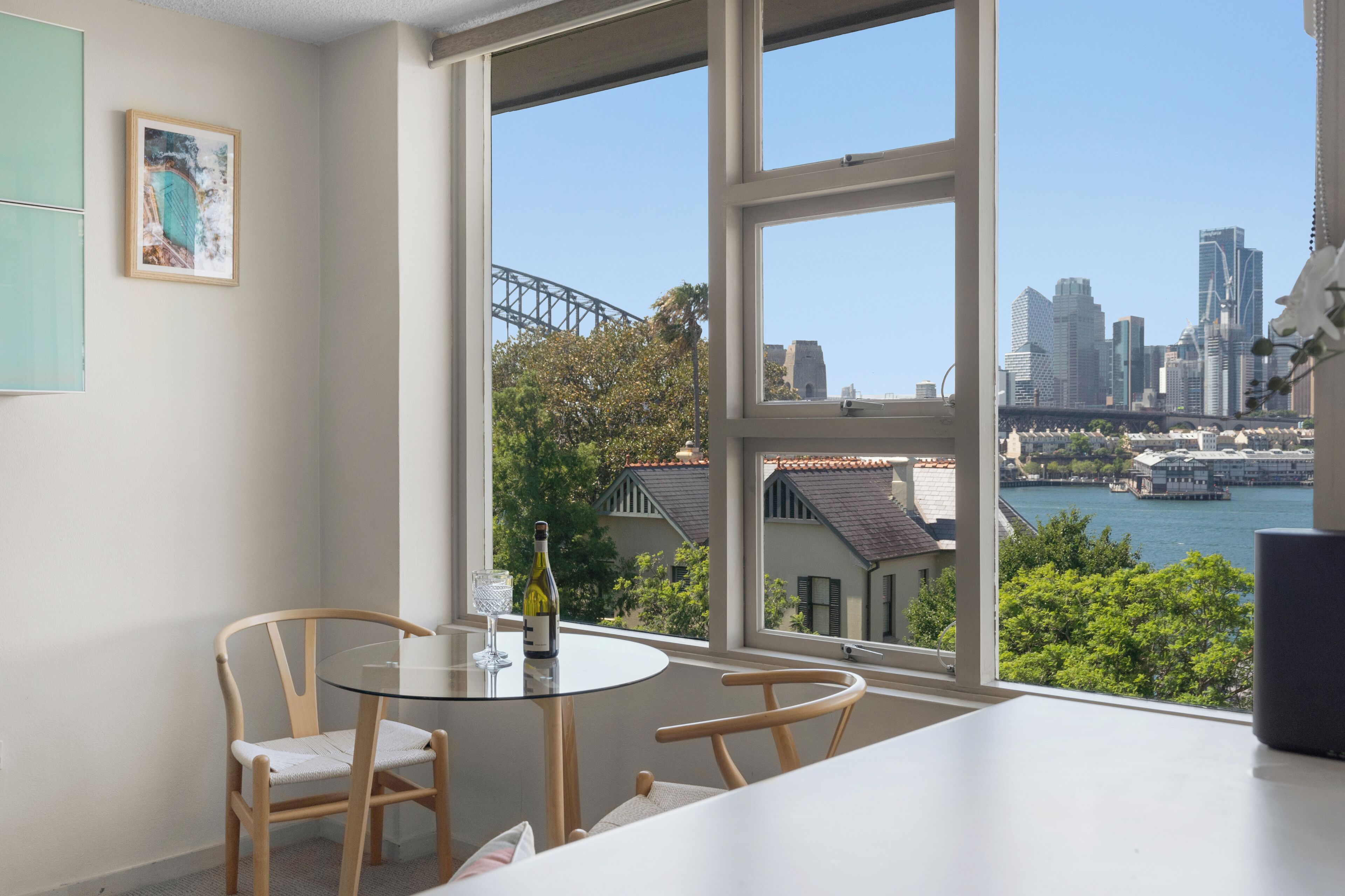 A dining area with a table and chairs, a framed picture on the wall, and a view of a city skyline and water.