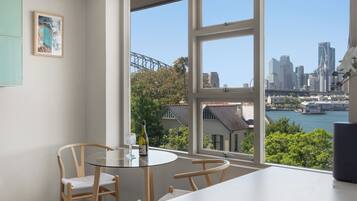 A dining area with a table and chairs, a framed picture on the wall, and a view of a city skyline and water.