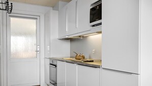 Une cuisine compacte avec des meubles blancs, un four encastré, un four à micro-ondes et un petit plan de travail sur lequel se trouve une bouilloire.