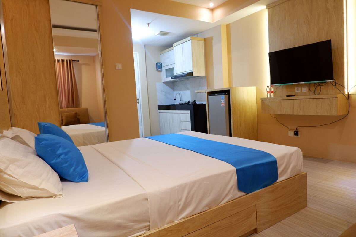 A hotel room with a bed, a TV, a small kitchenette, and a bathroom visible through an open door.