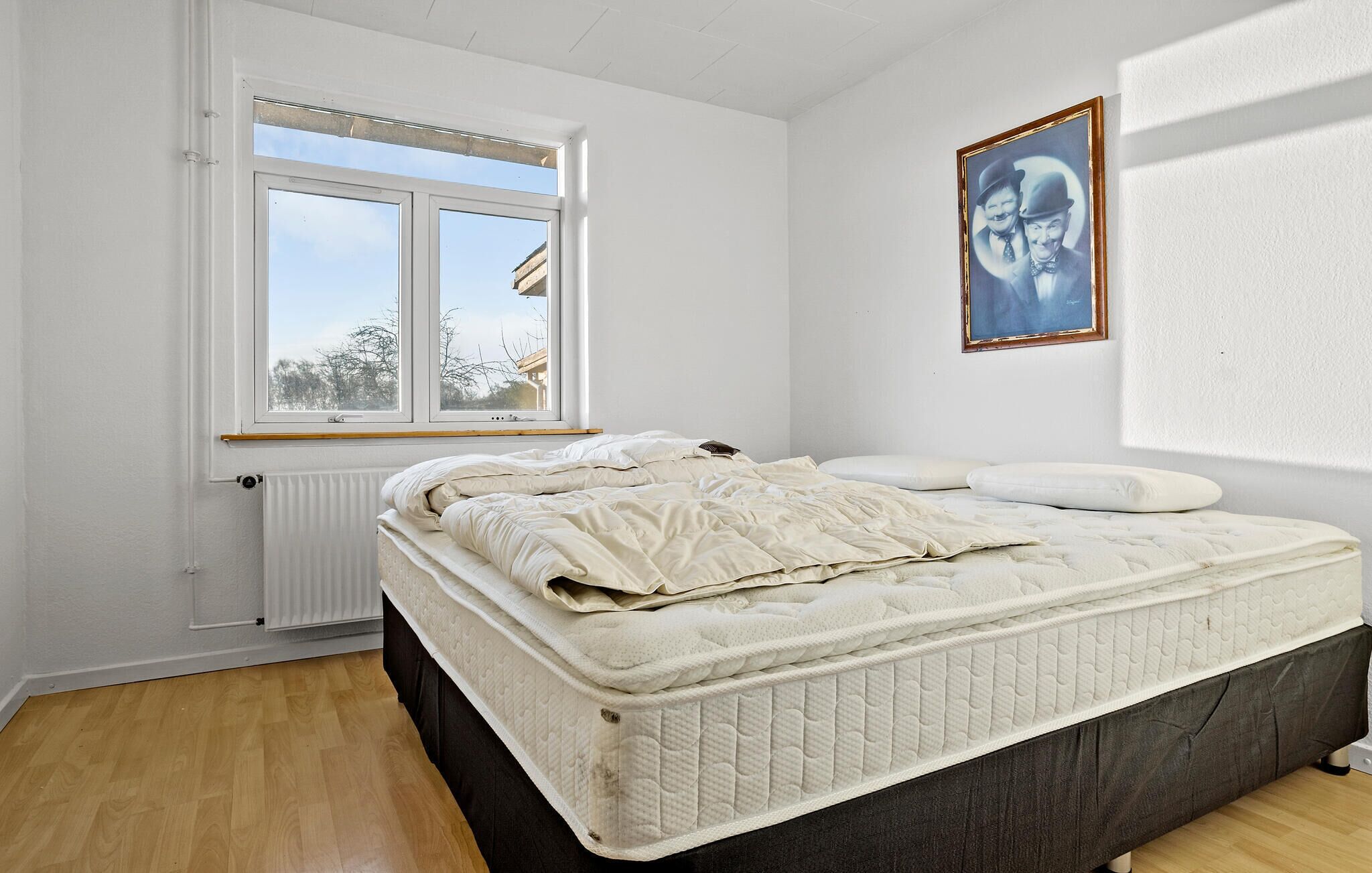 Un dormitorio con una cama, una ventana, un radiador y un cuadro en la pared.